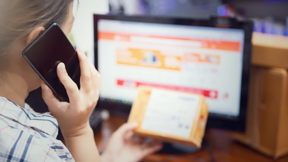 A shopper holding a parcel and a phone while an online store product page is open on a monitor behind her
