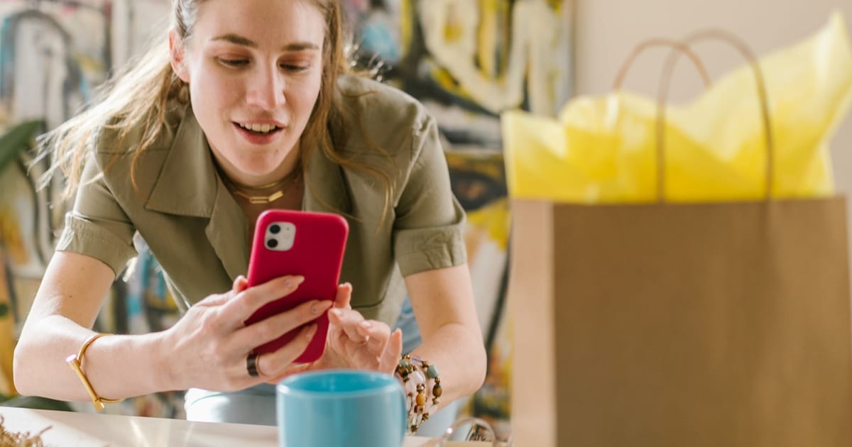 A shopper smiling at her phone while browsing an online store, with a shopping bag on the table beside her